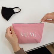 Woman zipping up The Pouch from Numi, a pink cotton canvas bag pictured on a desk with The Mask from Numi